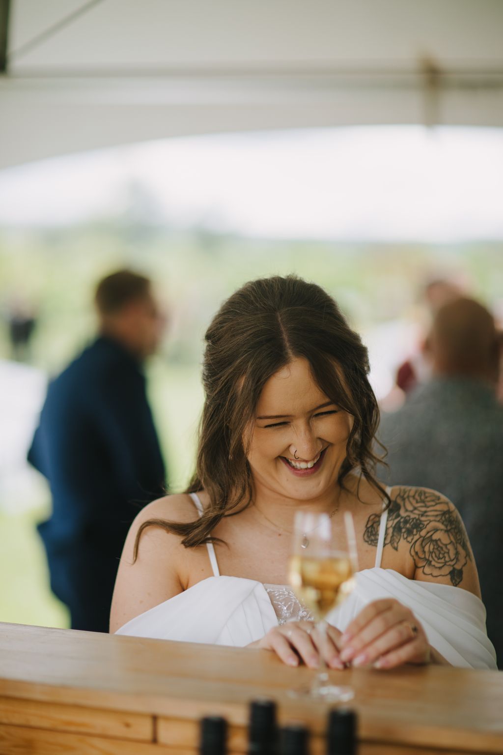 Smiling bride in white dress holding wine glass at outdoor reception, natural daylight filtering through tent