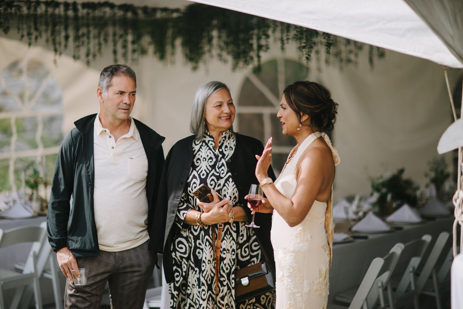 Two women in elegant attire smile while holding drinks at an indoor reception, with hanging greenery and soft natural light in the background