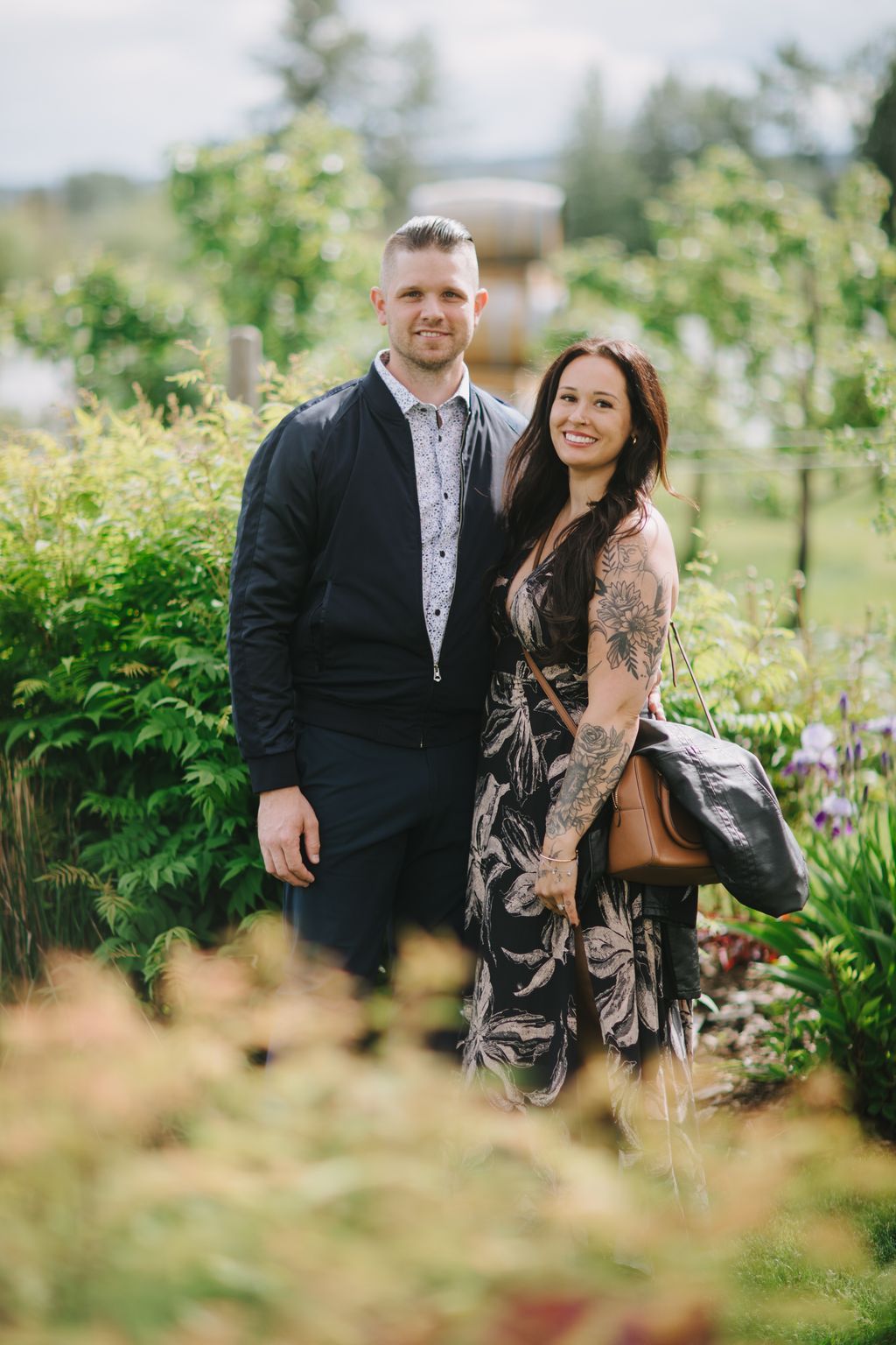 Couple smiling outdoors in a lush garden setting, man in navy jacket and patterned shirt beside woman in floral dress with tattoos