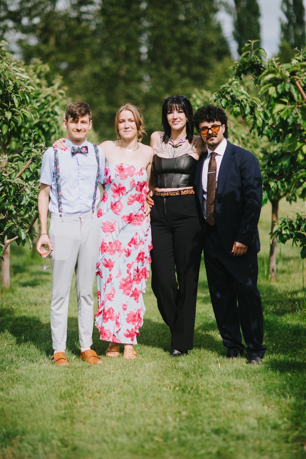 Four stylishly dressed guests stand together on a sunny lawn with blurred green trees behind them, smiling at the camera