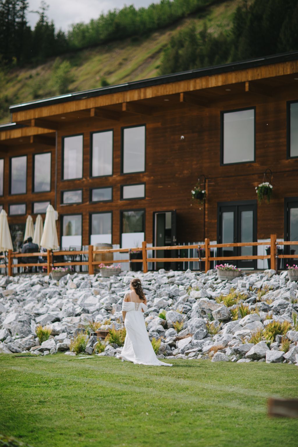 Bride in flowing white off-shoulder gown walks across manicured lawn toward modern wooden cabin nestled against forested hillside