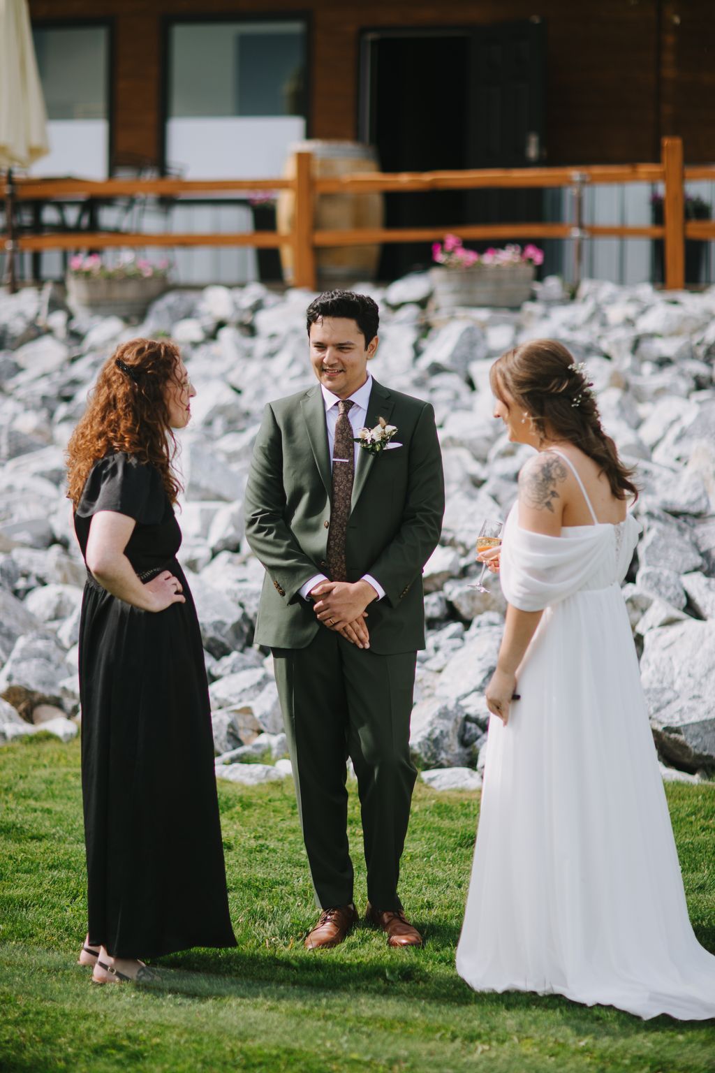 Groom in sage green suit with floral boutonniere smiles during outdoor wedding ceremony on grass with stone wall backdrop
