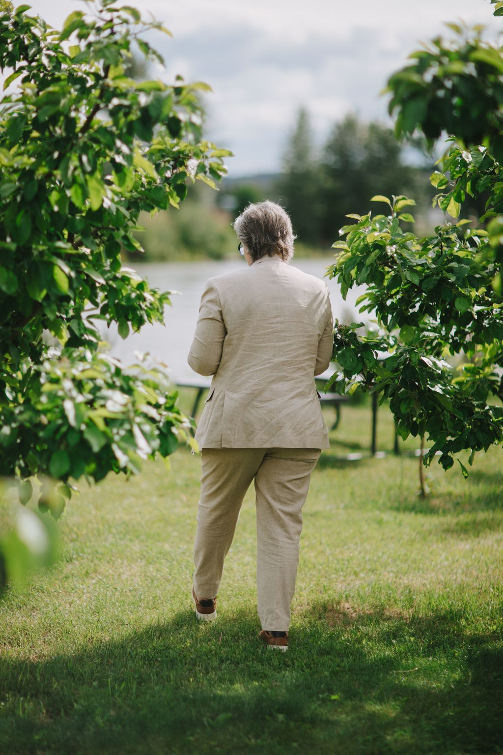 Older woman jogging through lush green gardens in cream linen suit, trees and water visible in soft-focused background