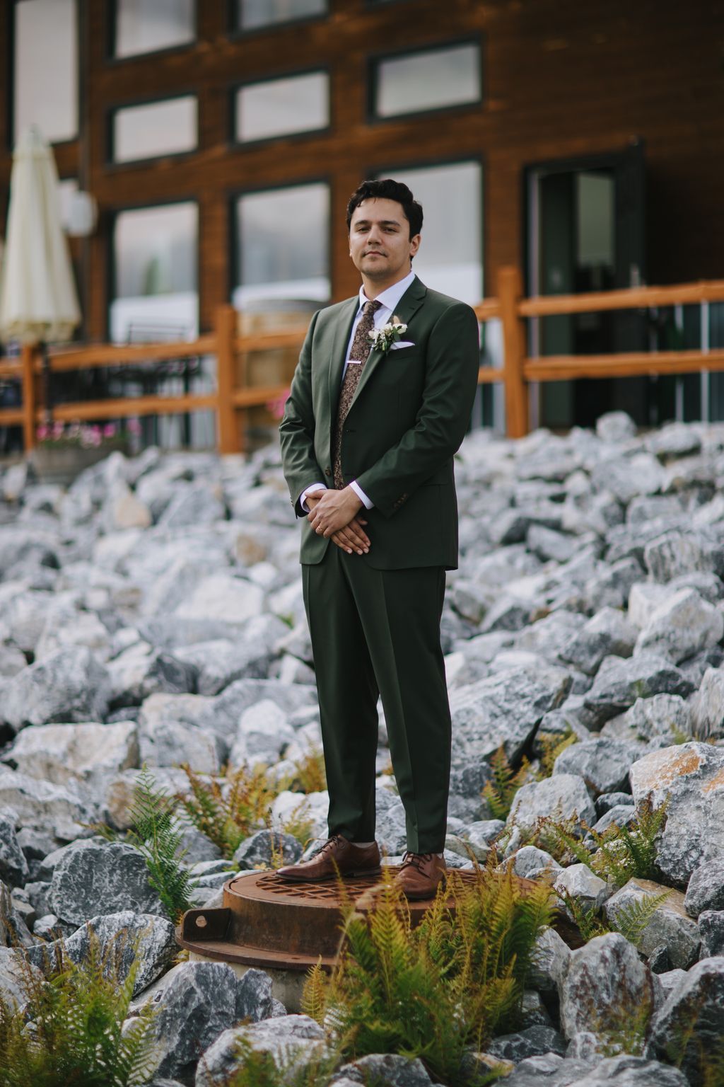 Groom in sage green suit and patterned tie stands on rocky terrain with wooden building backdrop