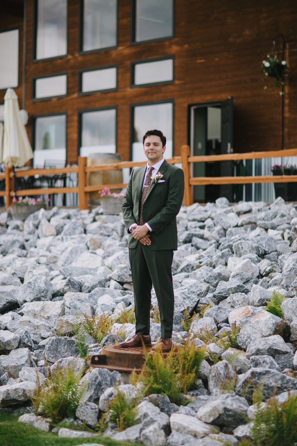 Groom in olive suit standing on rocky terrain with wooden cabin backdrop, wearing boutonniere and purple floral tie