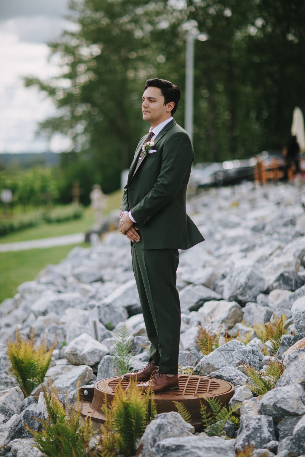 Groom in sage green suit standing on wooden platform among riverside rocks, gazing thoughtfully to the side with soft natural light filtering through trees b…