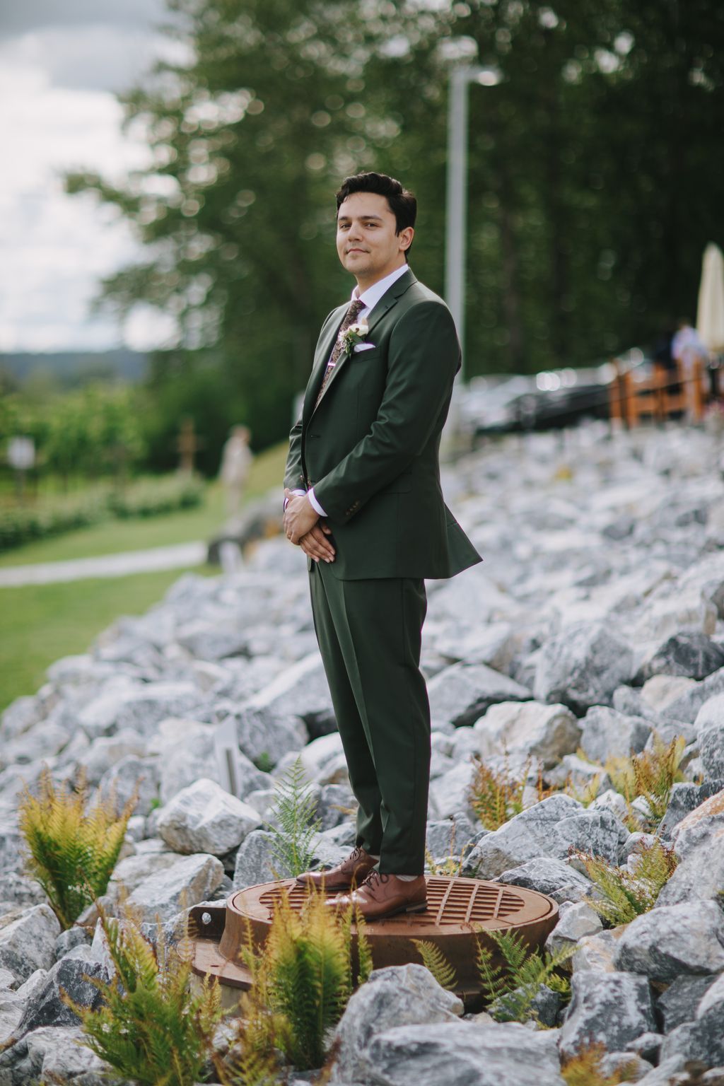 Groom in olive green suit standing on wooden platform among rocky terrain with ferns, smiling toward camera with blurred green foliage background