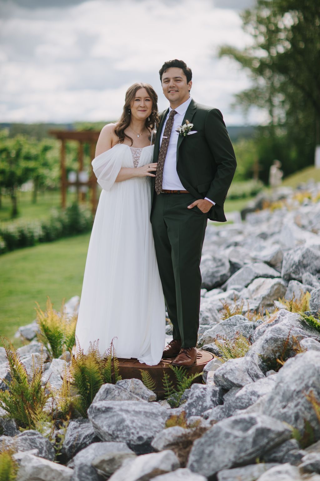 Bride in flowing white off-shoulder gown and groom in olive suit stand smiling on rocky landscape under cloudy sky