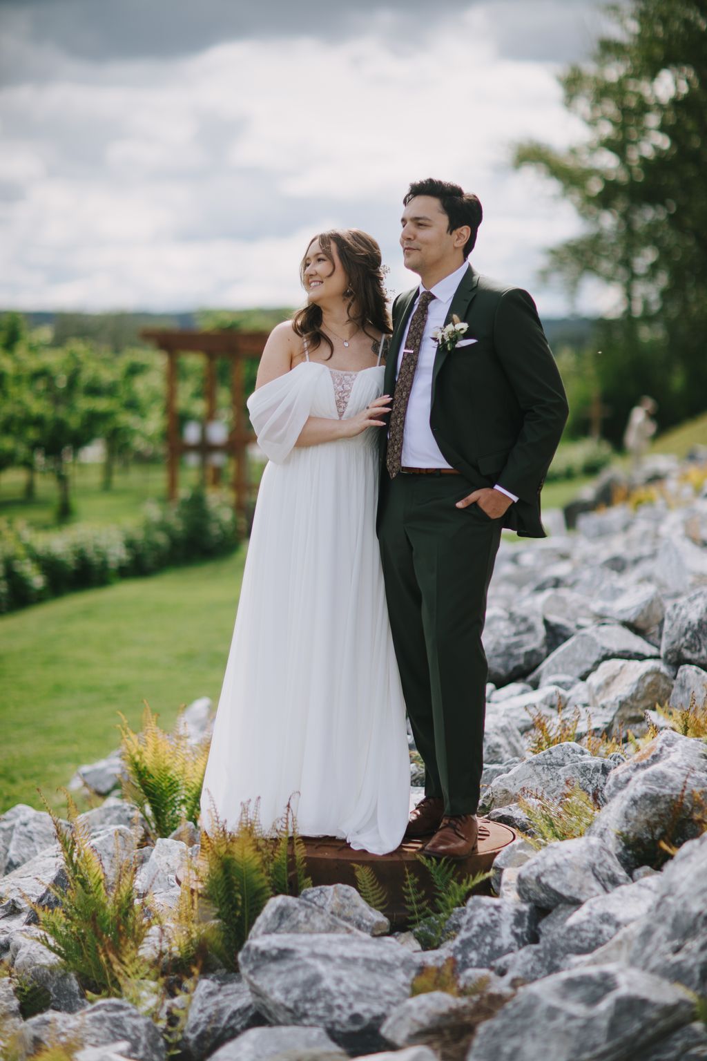 Bride in flowing white off-shoulder gown and groom in dark suit stand together on rocky outcrop overlooking pastoral landscape under cloudy sky