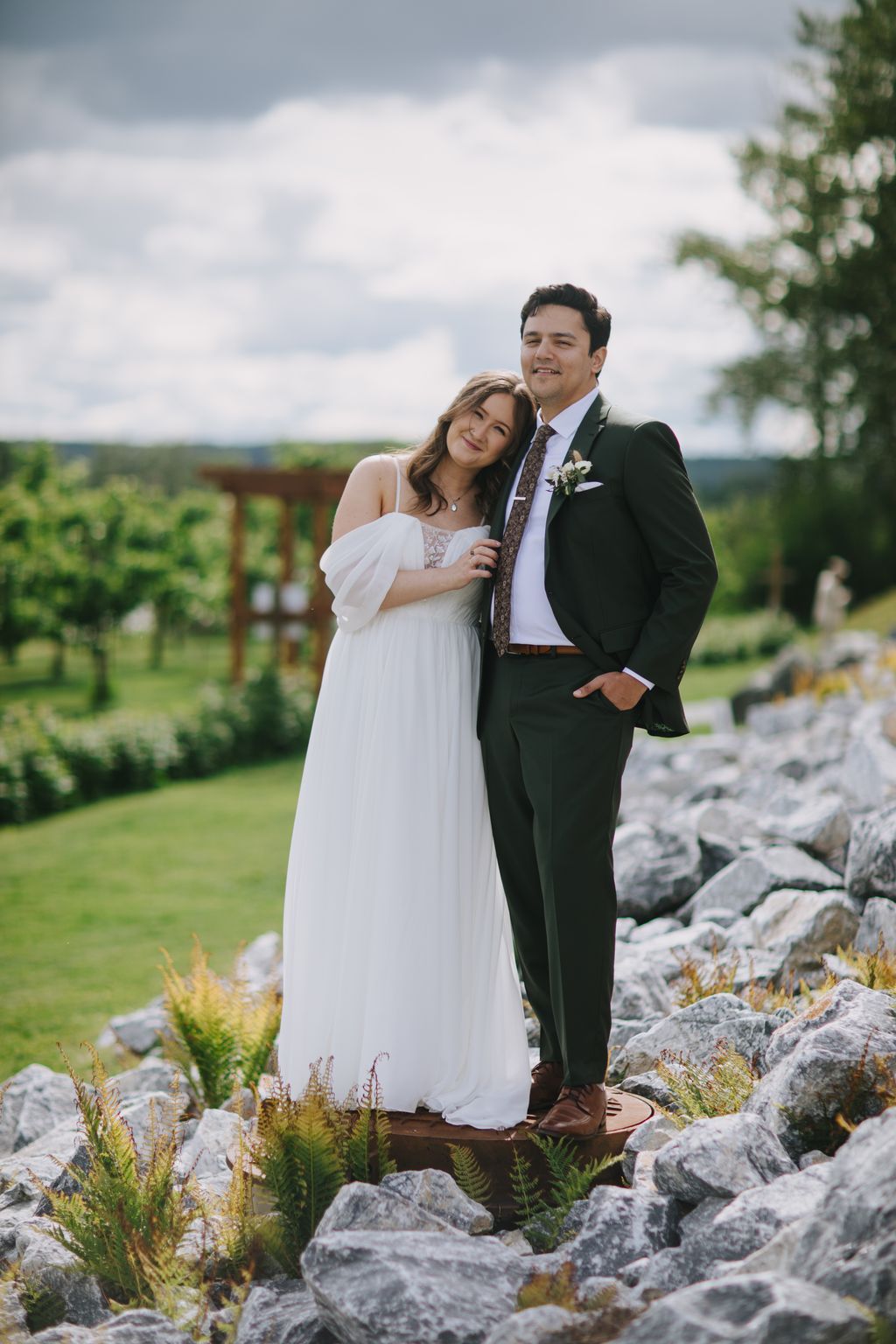 Bride in flowing white off-shoulder gown and groom in dark suit stand together on stone steps with rolling fields and cloudy sky behind them