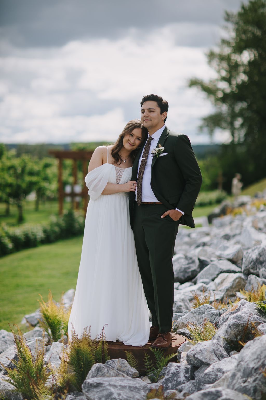 Bride in flowing white off-shoulder gown and groom in dark suit embrace on rocky outcrop overlooking pastoral landscape under dramatic cloudy sky