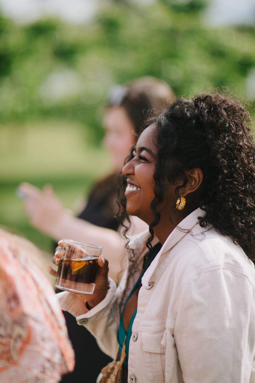 Woman in cream jacket holding a drink, smiling outdoors with blurred green background and guests behind her