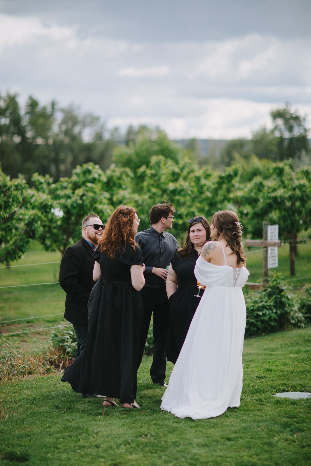 Bride in white dress and wedding party in black attire stand together on green lawn with forested hillside backdrop under cloudy sky