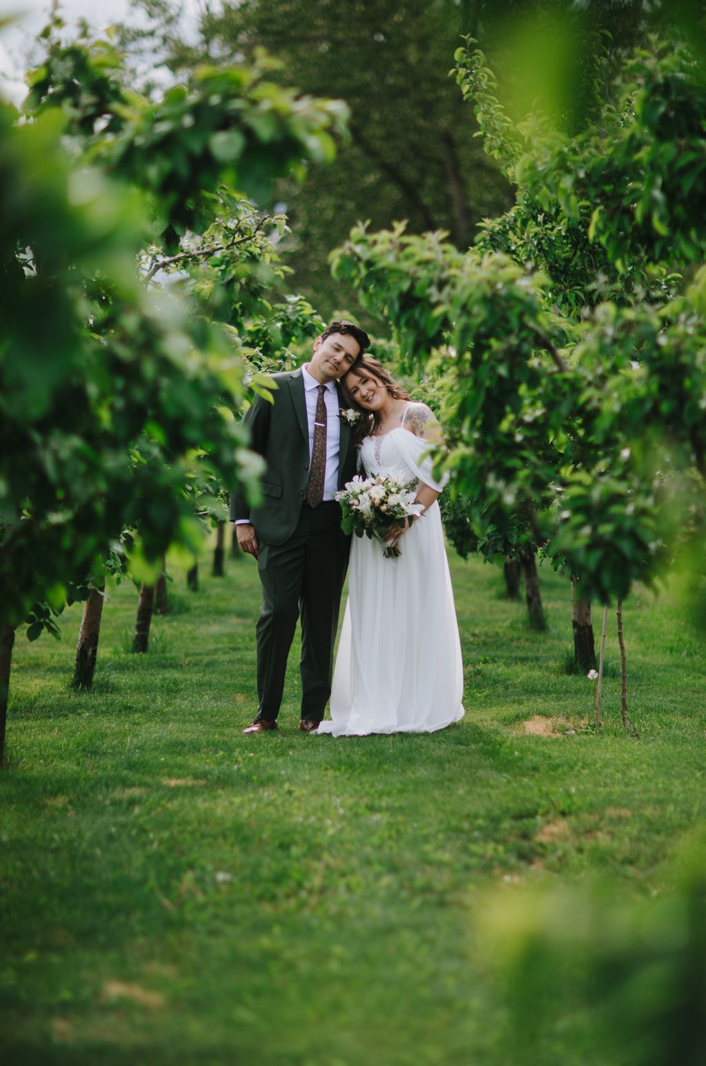 Bride and groom embrace among lush green foliage in a garden, soft natural light filtering through leaves
