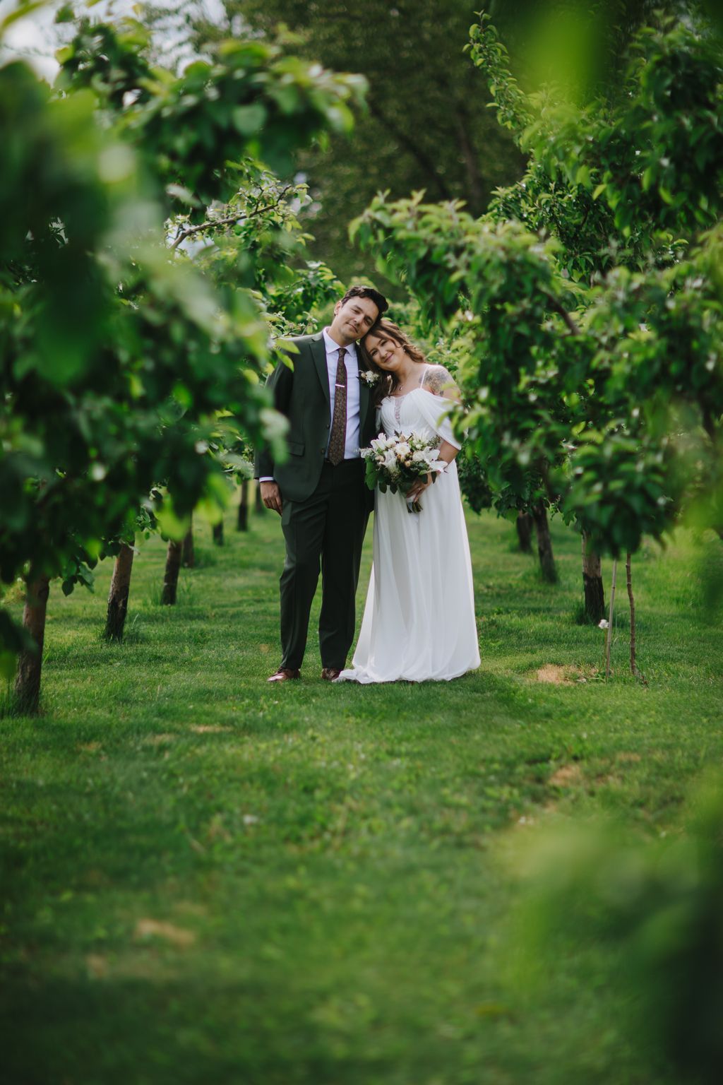 Bride and groom stand together in lush green garden surrounded by leafy branches, bride holding white bouquet, soft natural light filtering through foliage