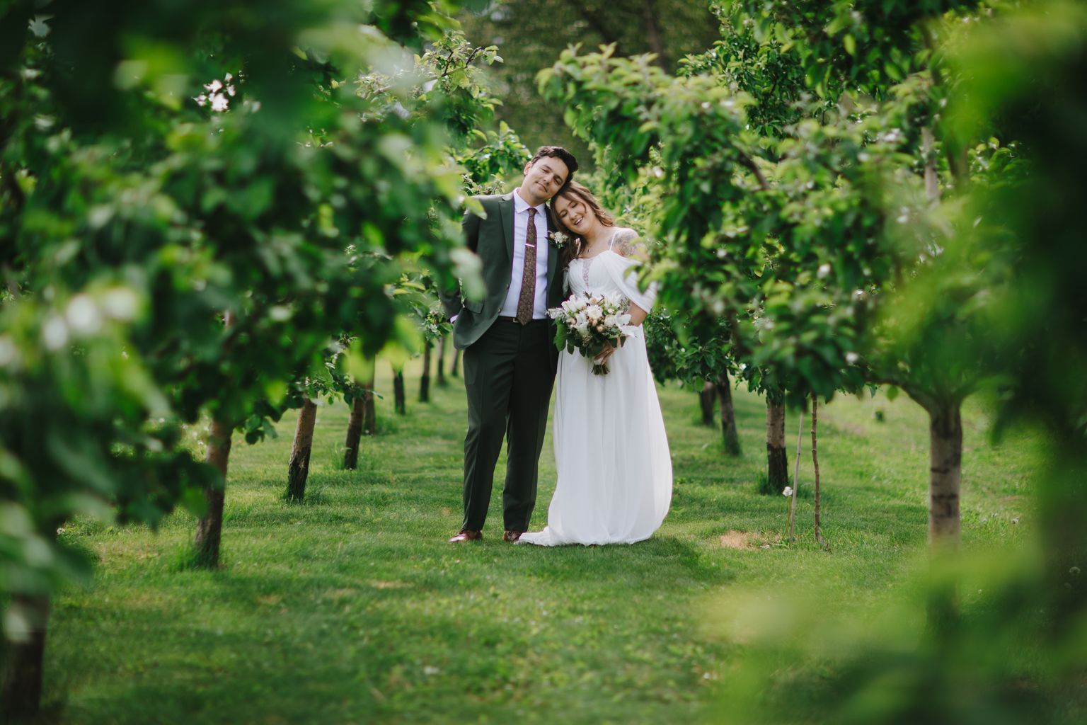 Bride and groom embrace beneath lush green vines in a sunlit garden, holding a white bouquet