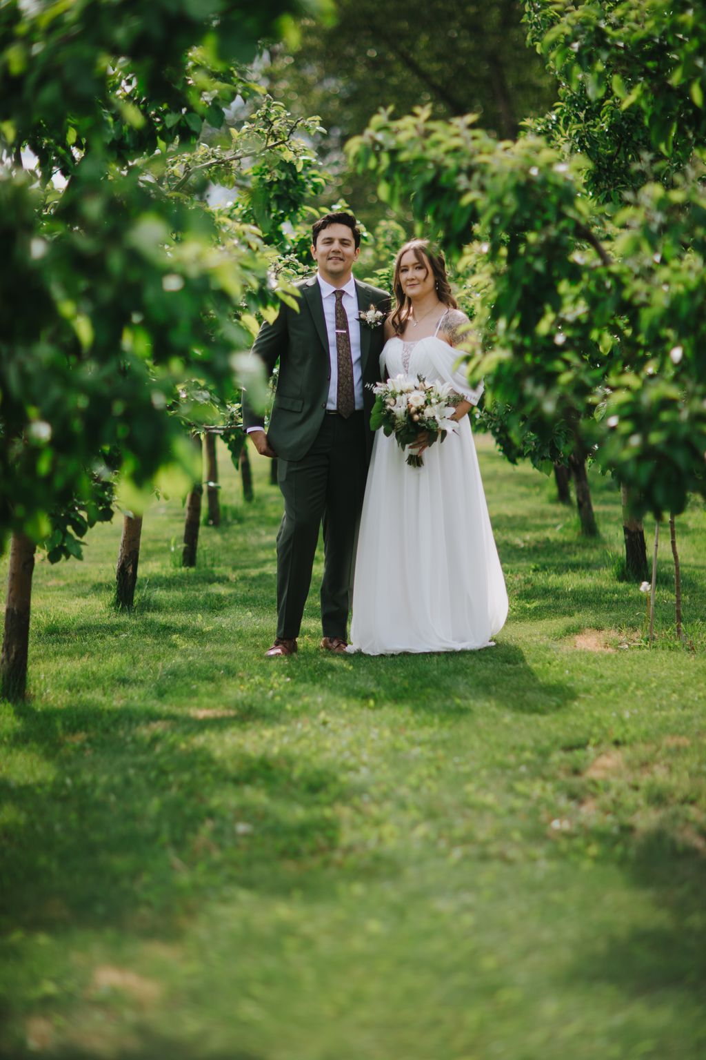 Bride and groom stand beneath flowering tree branches in lush garden, bride holding white bouquet, soft natural light filtering through green foliage