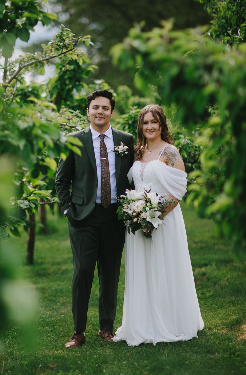 Bride and groom stand together on green lawn with lush garden backdrop, bride in flowing white dress holding white bouquet, groom in dark suit with patterned…