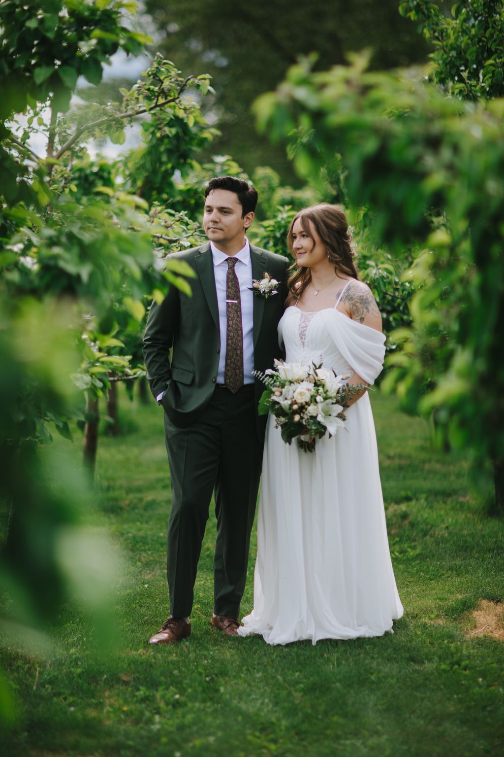 Bride in flowing white gown and groom in dark suit stand together on green lawn surrounded by lush foliage, holding white flower bouquet