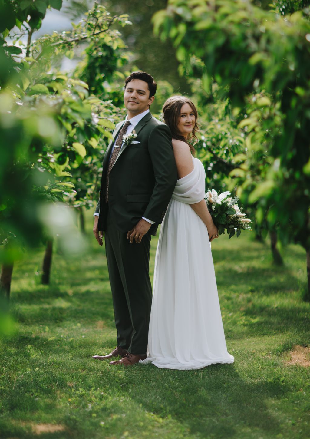 Bride in white off-shoulder gown and groom in dark suit stand together on grass with lush green foliage behind them