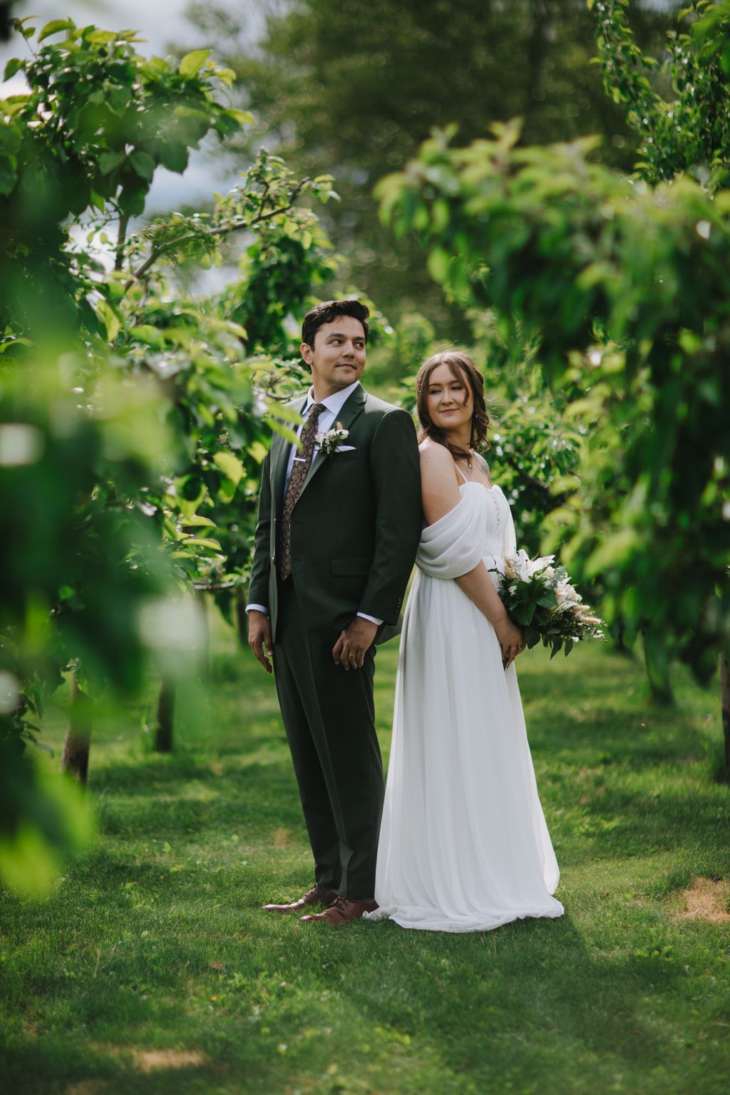 Groom in charcoal suit and bride in draped white gown stand together on green lawn surrounded by lush foliage, both gazing upward in soft natural light