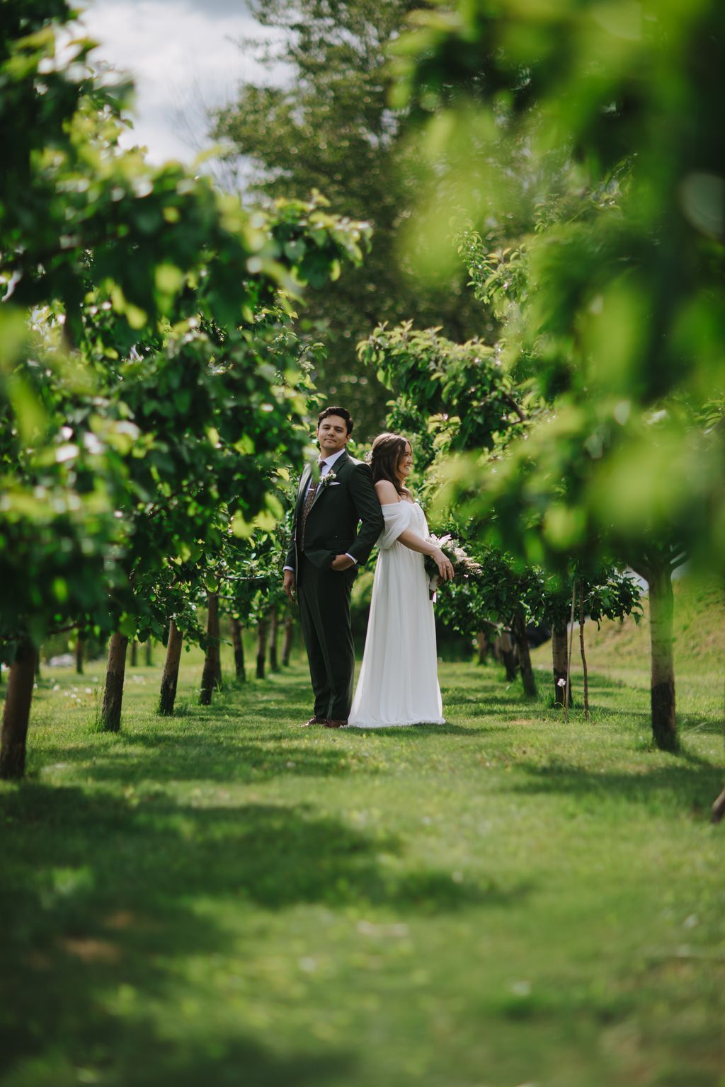 Bride and groom stand together in a lush green vineyard, soft natural light filtering through leafy vines framing the couple