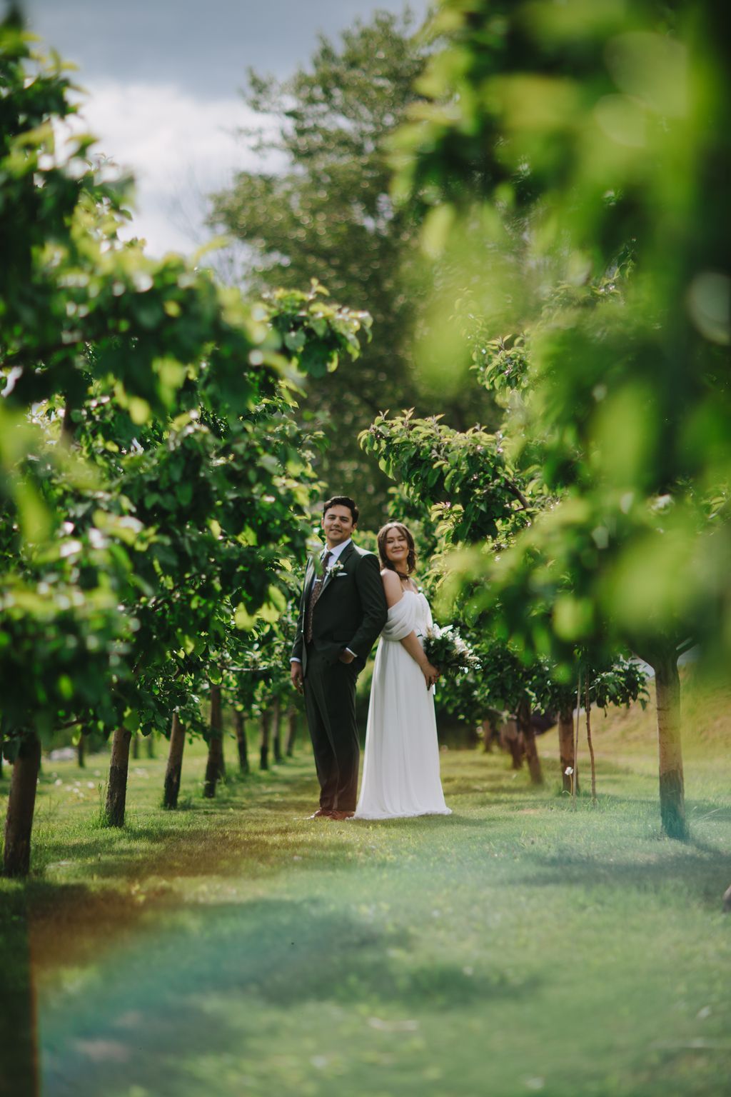 Bride in white off-shoulder gown and groom in dark suit stand together in lush vineyard rows under bright daylight