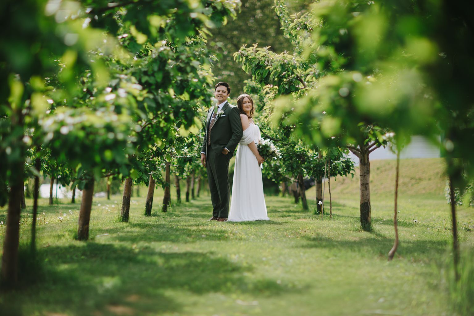 Bride and groom standing together in a lush vineyard, surrounded by green foliage, smiling at the camera in soft natural light