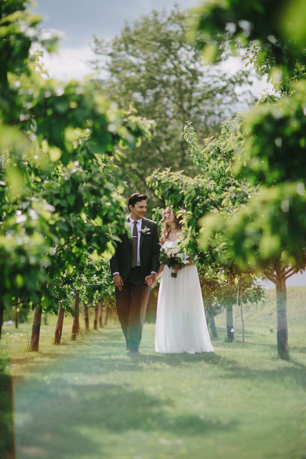 Bride and groom walking hand-in-hand through a blooming orchard lined with flowering trees on a sunny day
