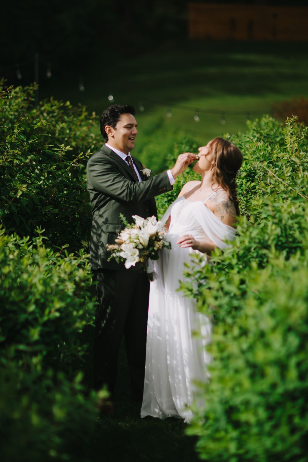 Groom in olive suit feeds bride in flowing white dress during evening garden ceremony surrounded by lush green hedges