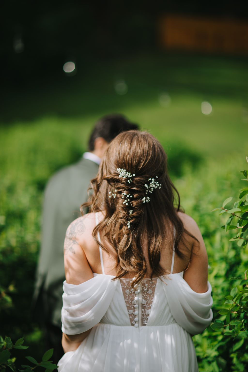 Bride from behind wearing white dress with baby''s breath woven through braided hair, soft green garden backdrop