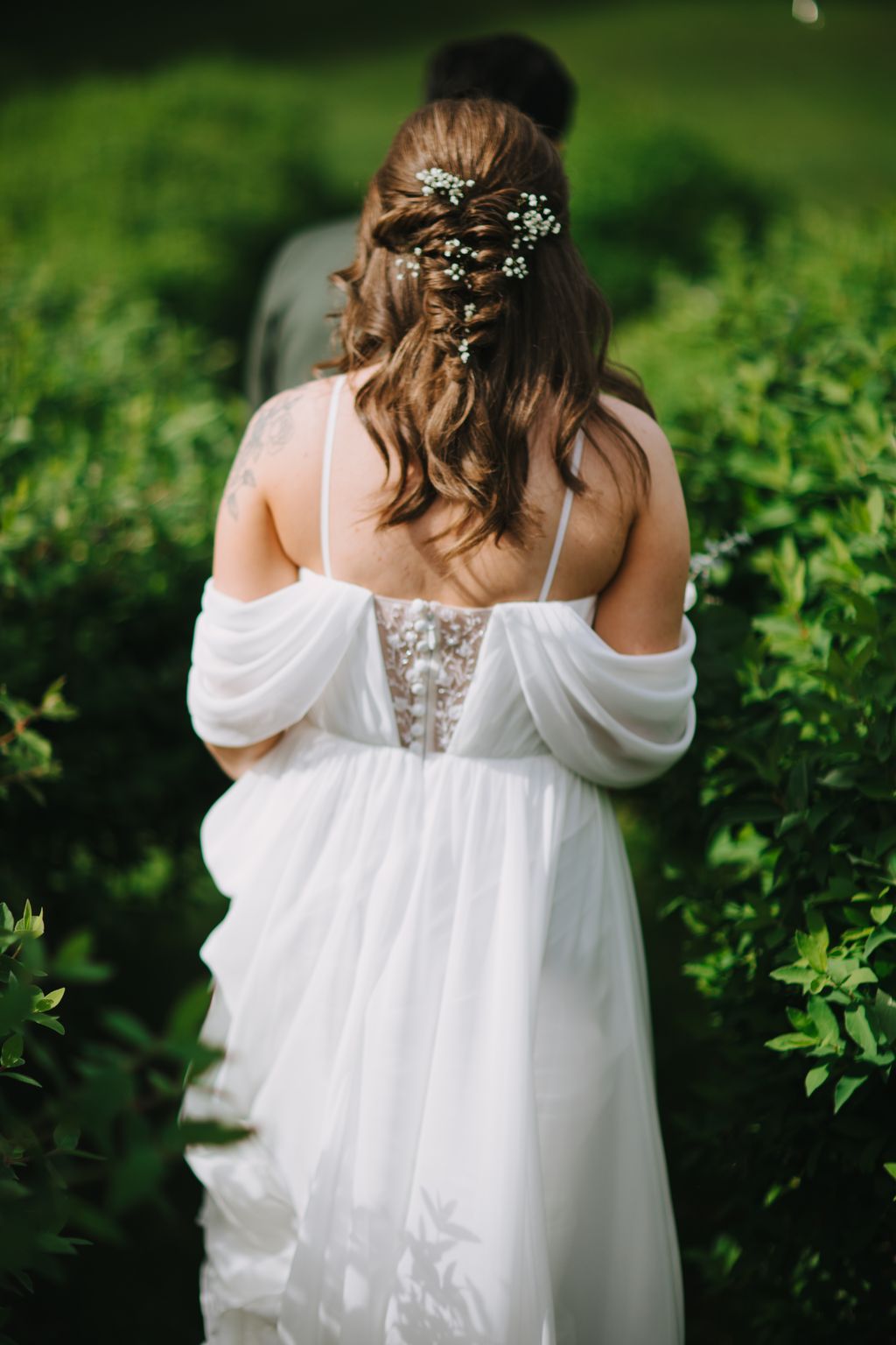 Bride in flowing white gown with baby''s breath woven through half-up hair, viewed from behind against soft green garden background