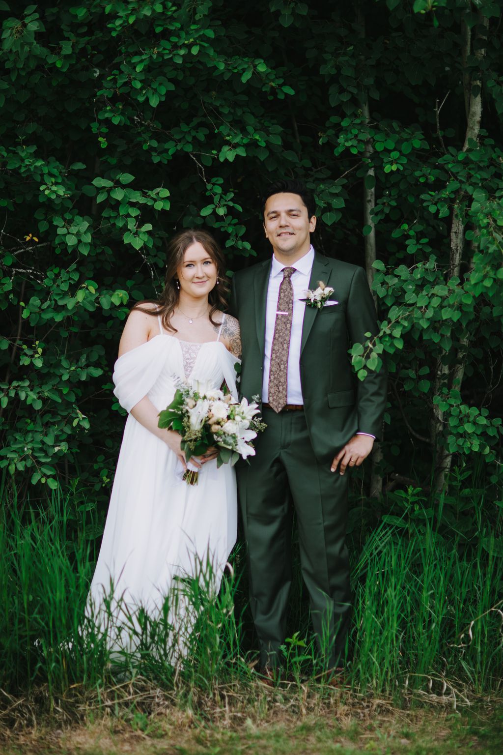 Bride in white dress and groom in forest green suit stand together against lush green foliage backdrop, holding white bouquet