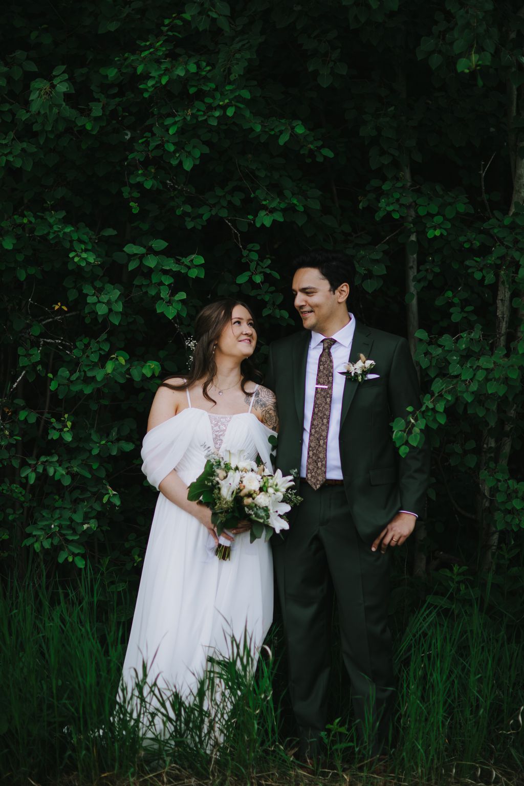 Bride in white off-shoulder dress and groom in dark suit smile together against lush green foliage backdrop, holding white flower bouquet