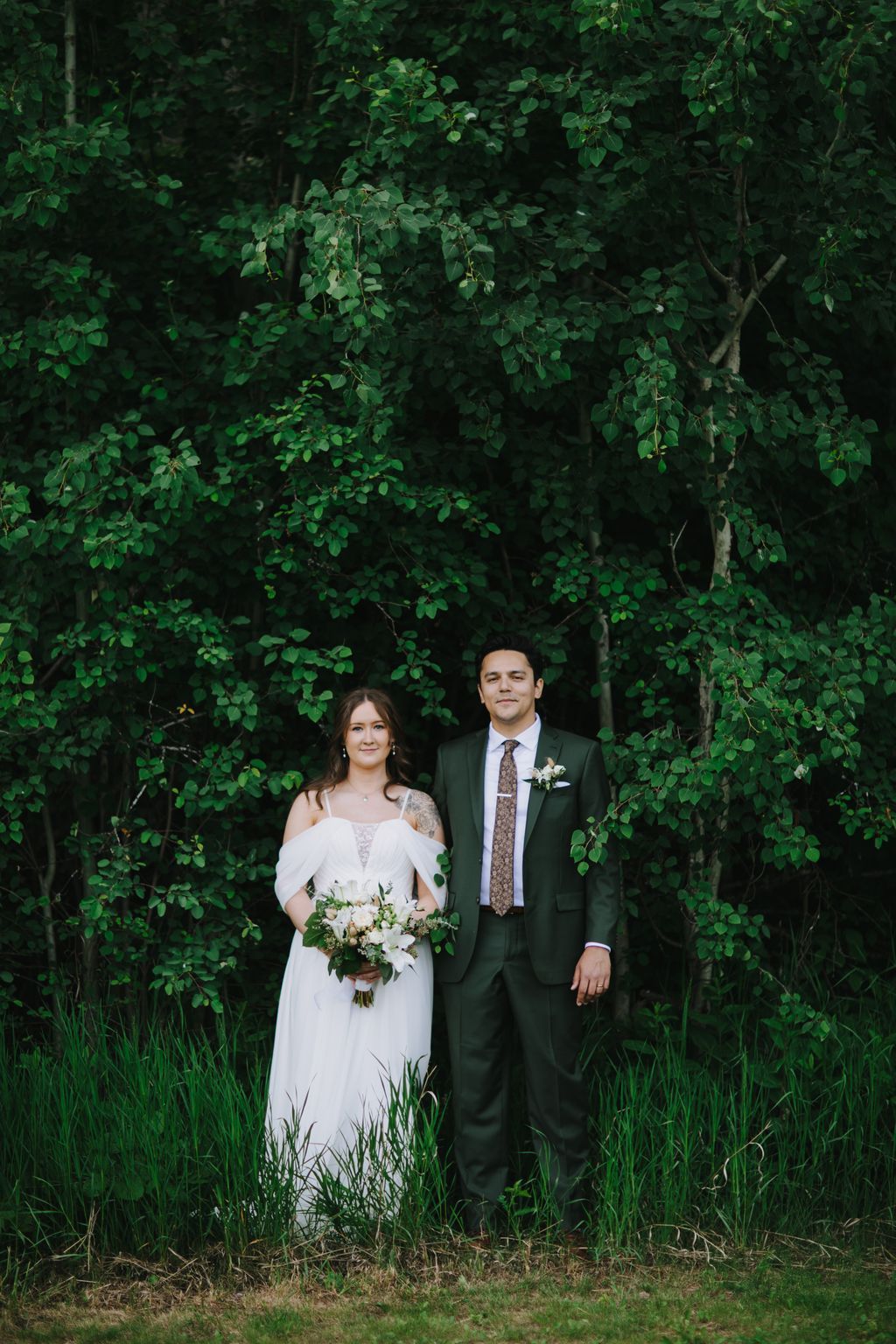 Bride in off-shoulder white gown and groom in sage green suit stand before lush green foliage backdrop