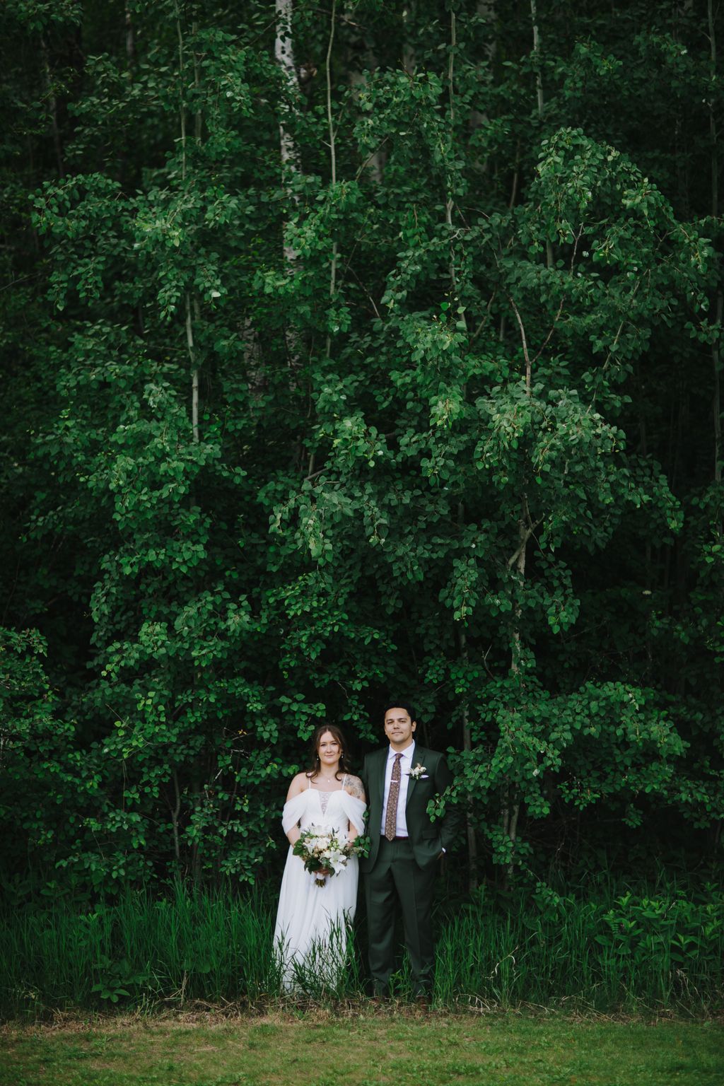 Bride in white off-shoulder gown and groom in charcoal suit stand before lush green foliage backdrop, holding wedding bouquet