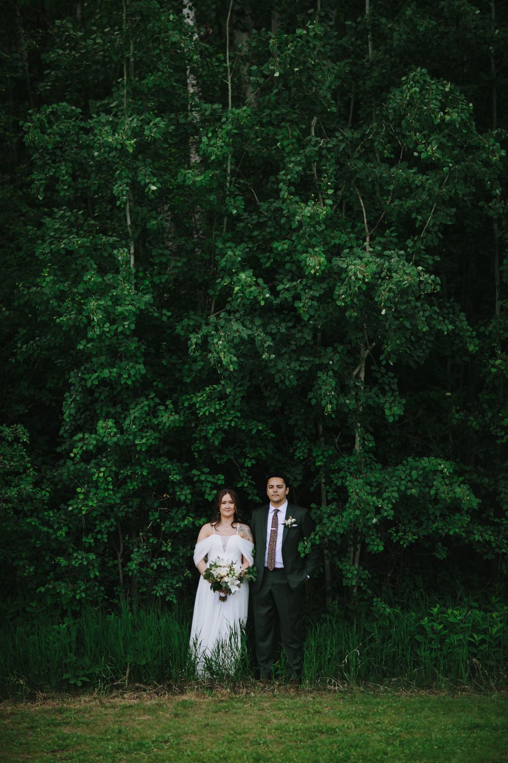 Bride and groom stand against lush green foliage, bride holding white bouquet, natural outdoor garden setting