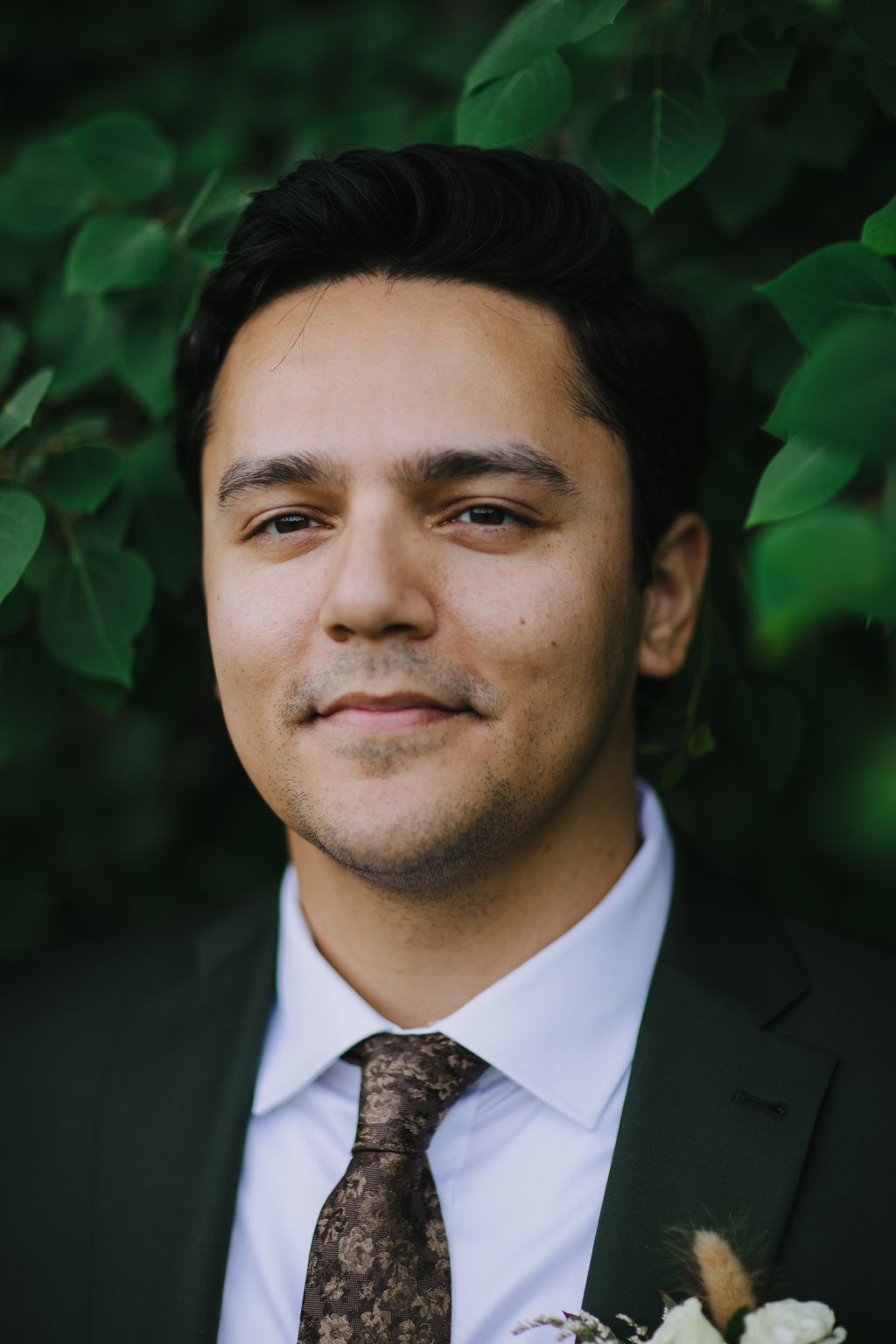 Groom in white dress shirt and patterned brown tie against soft green foliage backdrop, gentle natural light