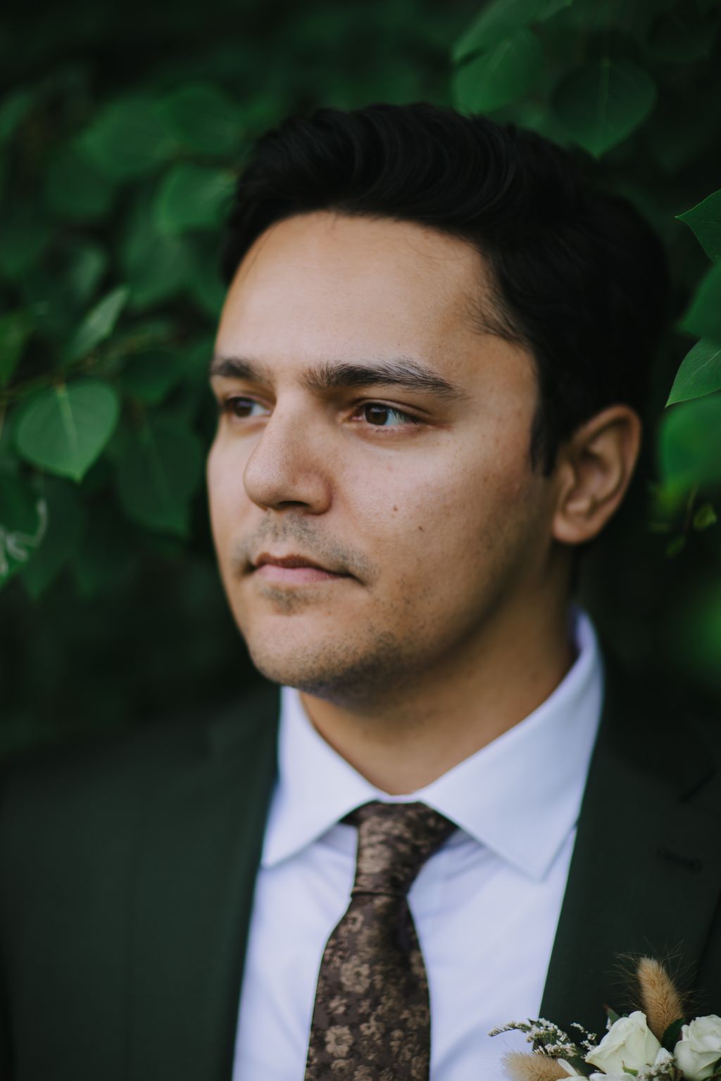 Groom in white dress shirt and patterned tie gazes thoughtfully away, green foliage soft-focused behind him