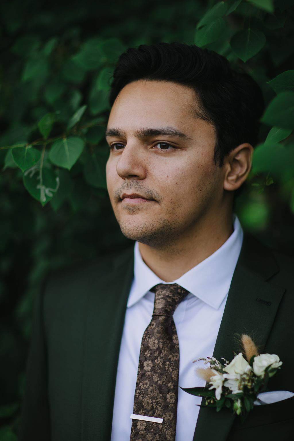Groom in light blue shirt and patterned brown tie with boutonniere against green foliage backdrop, looking thoughtfully to the side