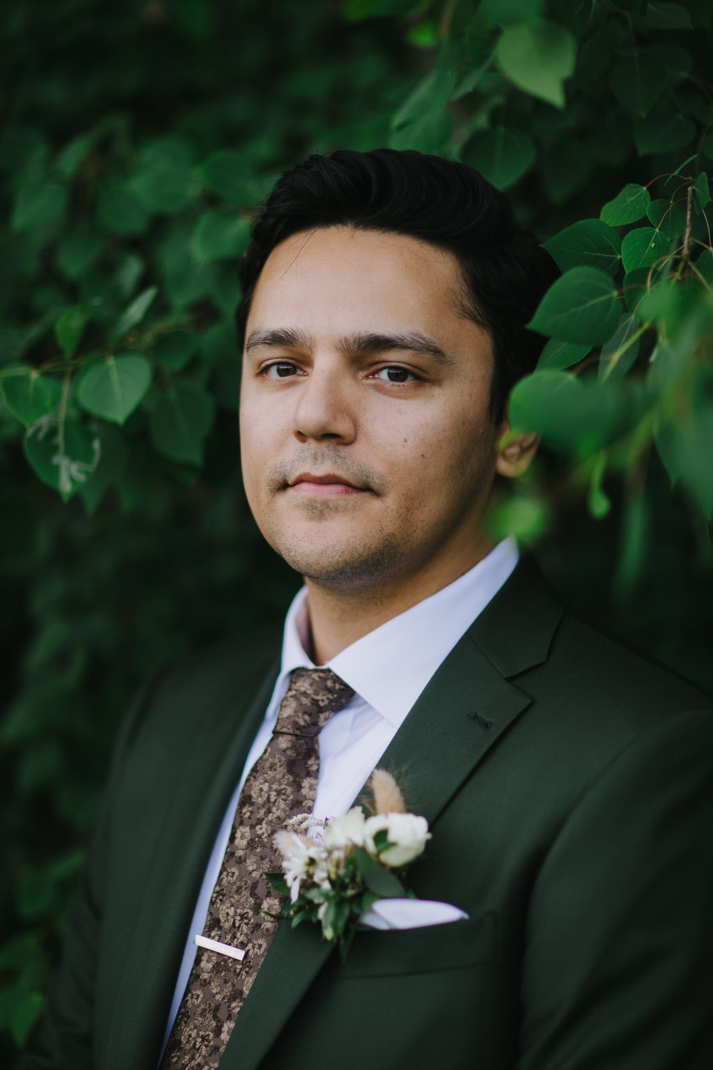 Groom in dark suit and patterned tie with white boutonnière against lush green foliage backdrop