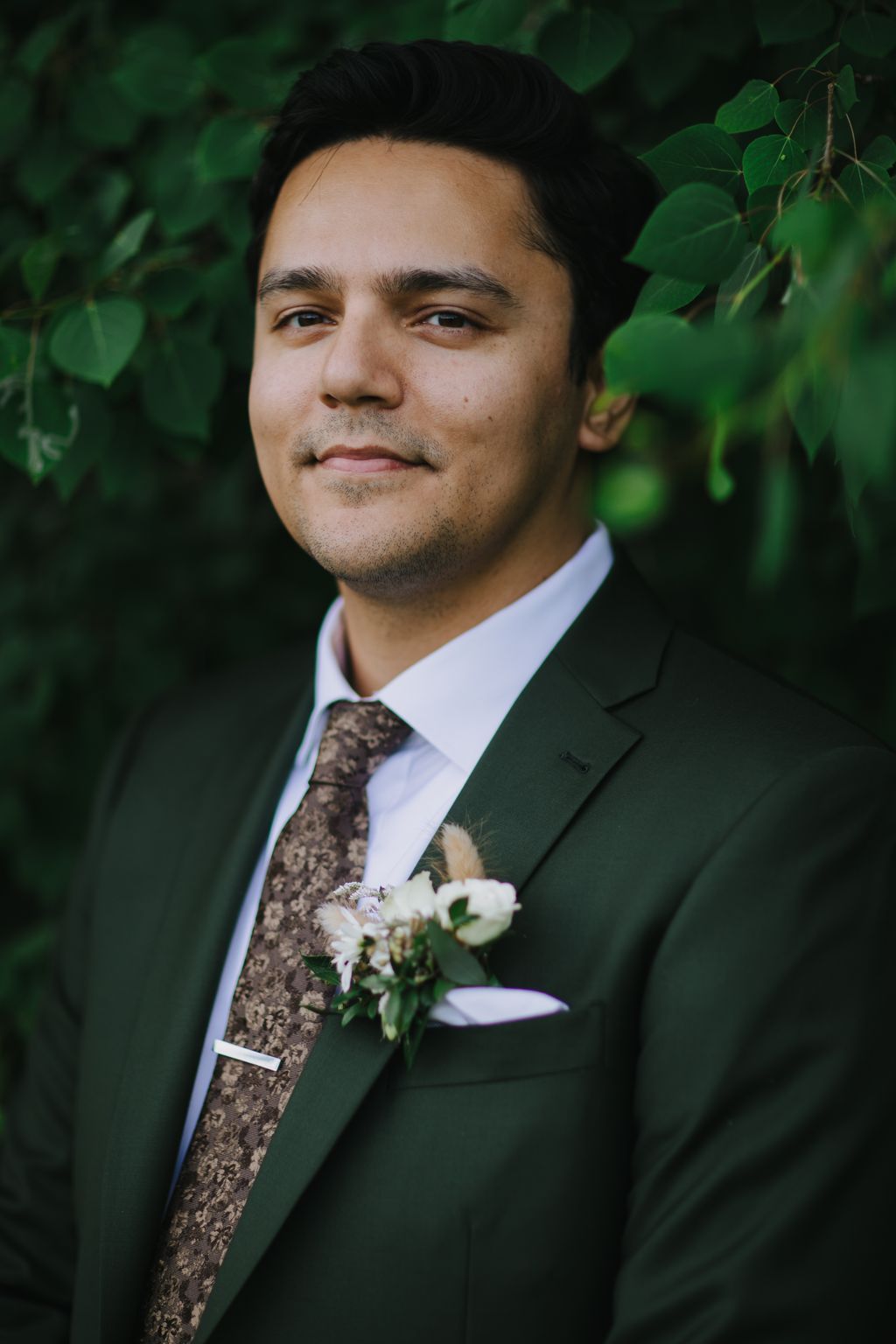 Groom in dark green suit with patterned tie and white boutonniere against soft green foliage backdrop