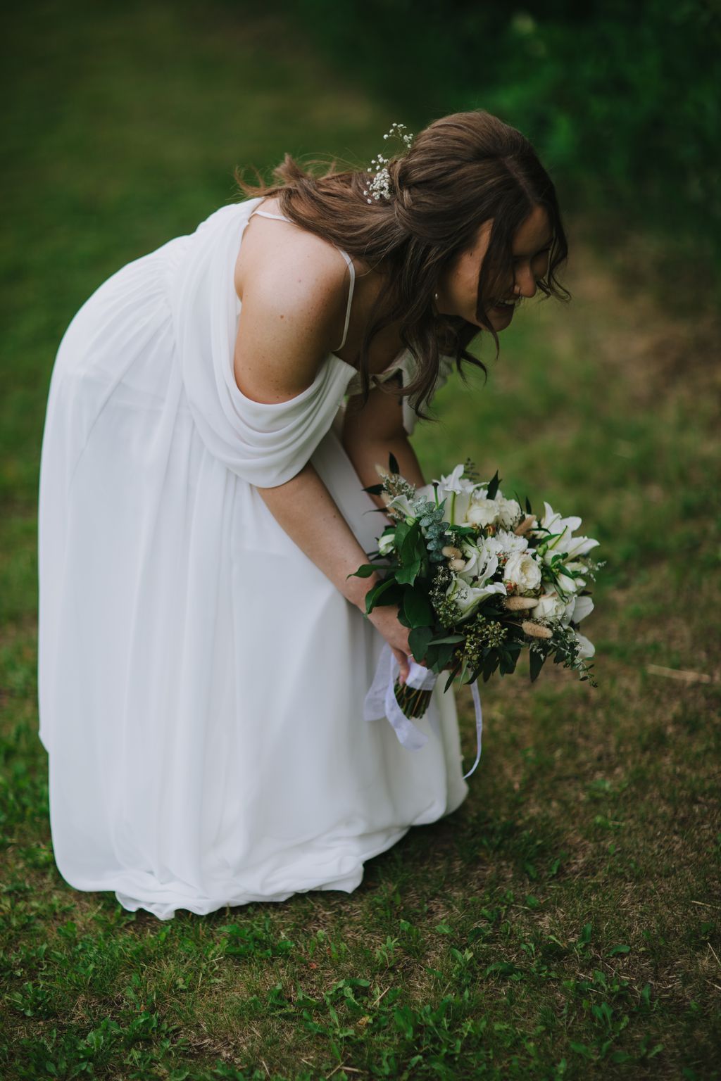 Bride in white dress holding white and green bouquet, looking down with baby''s breath in upswept hair, standing on grass with soft green background