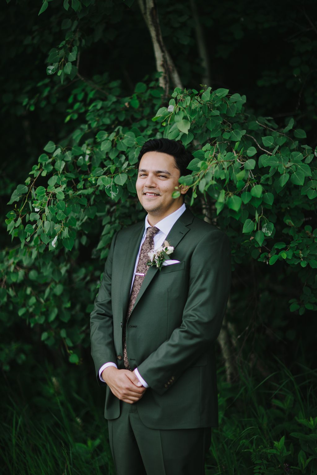 Groom in sage green suit with floral tie and boutonniere smiles against lush green foliage backdrop
