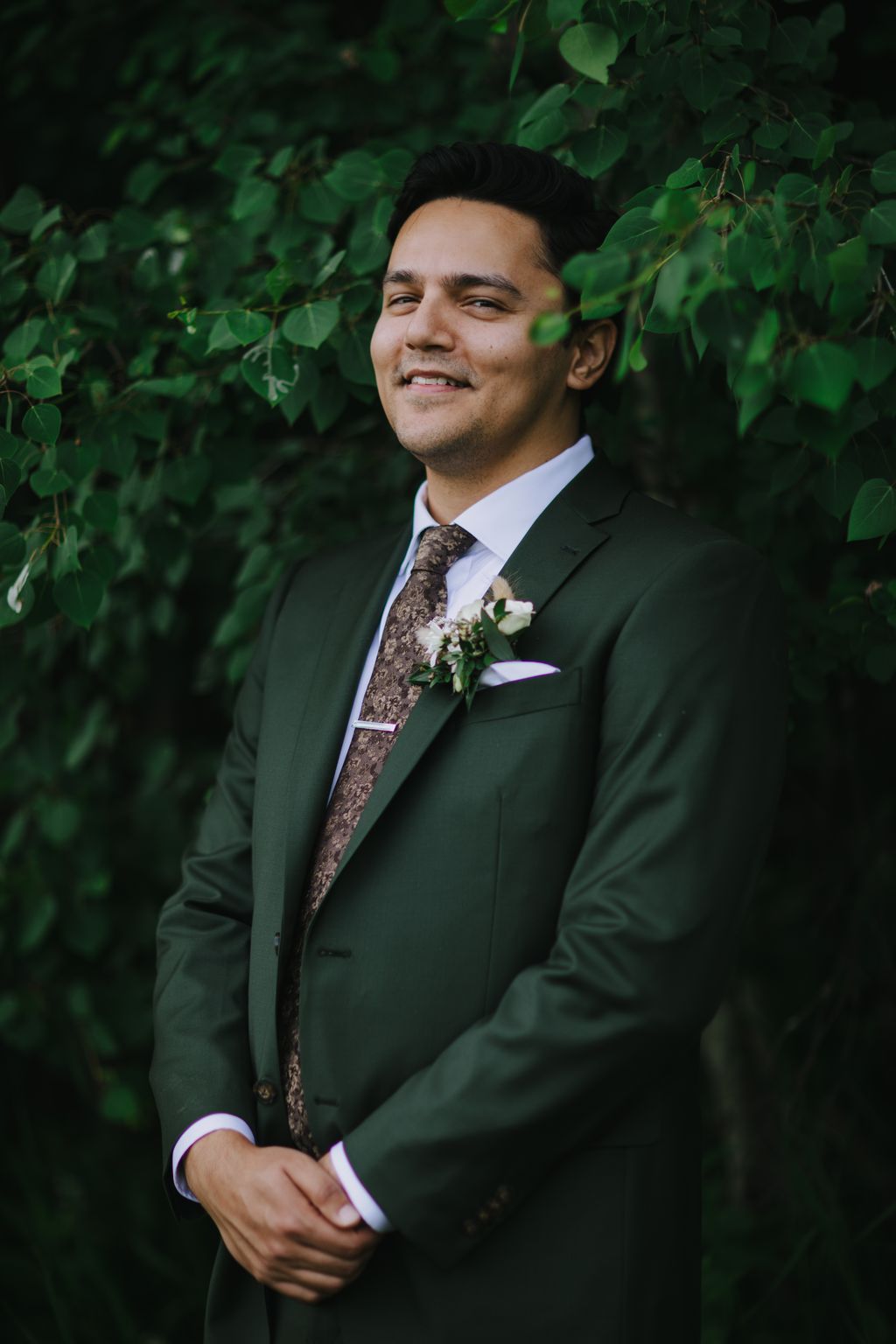 Groom in forest green suit with patterned tie and boutonniere smiles against lush green foliage backdrop