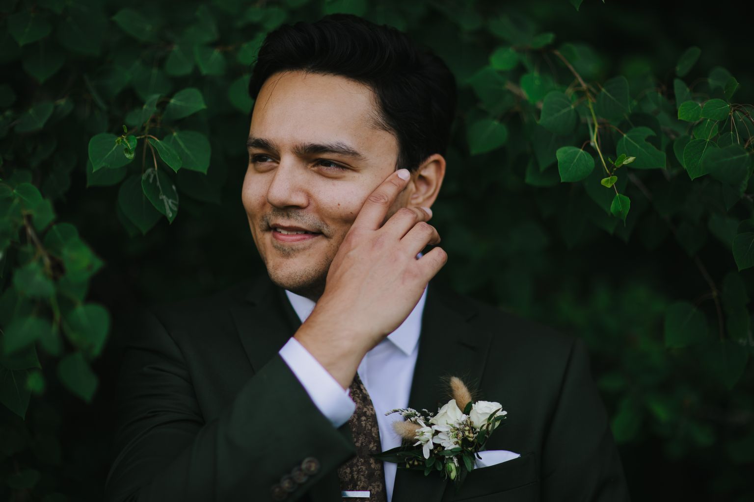 Groom in dark suit and patterned tie with boutonniere gazes downward thoughtfully against soft green foliage background