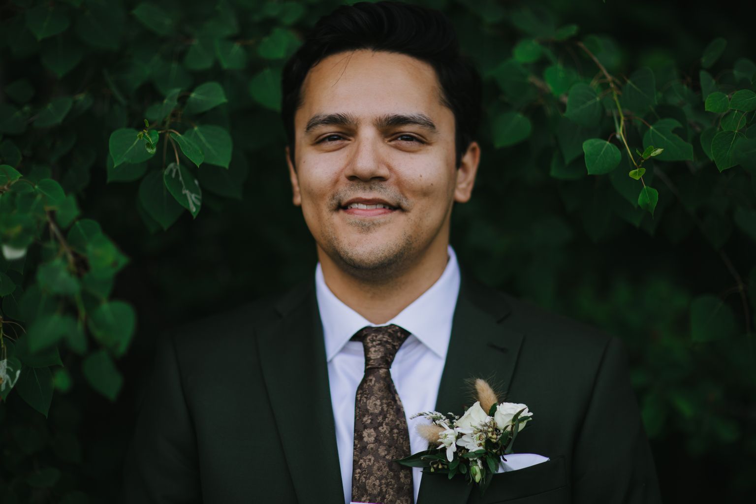 Groom in dark suit and patterned brown tie with white boutonniere, smiling at camera against blurred green background