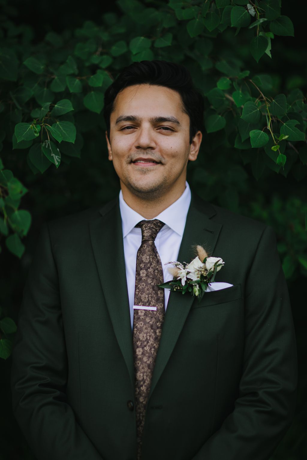 Groom in dark green suit with patterned tie and white boutonniere smiling at camera against lush green foliage backdrop