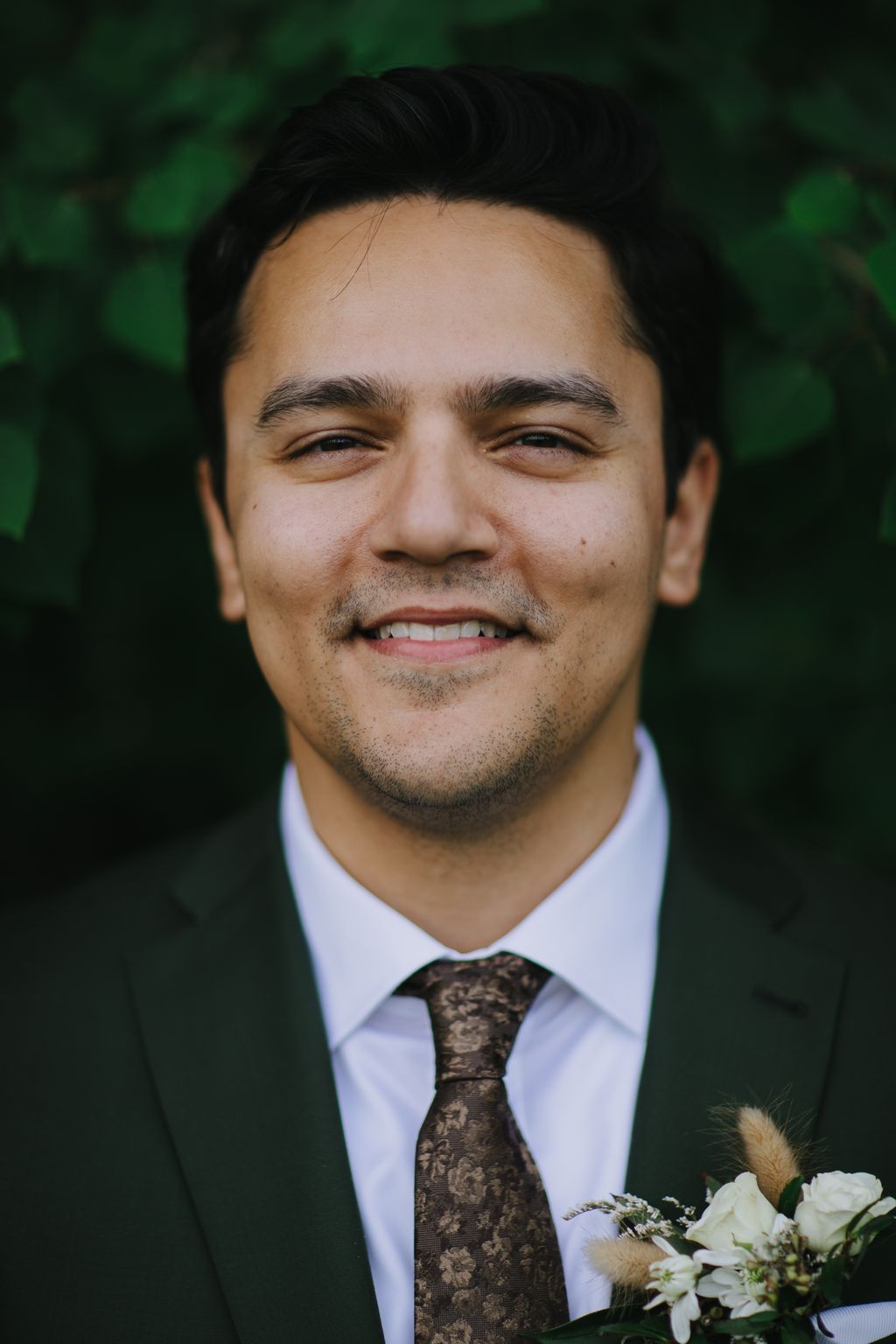 Groom in white dress shirt and patterned brown tie smiling at camera with boutonniere, green blurred background