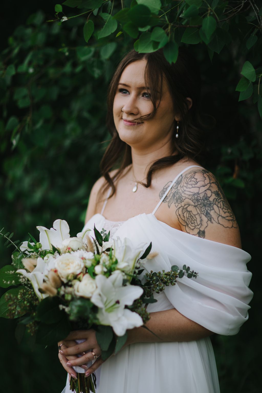 Bride in white off-shoulder dress holding white bouquet against lush green foliage backdrop, smiling softly at camera